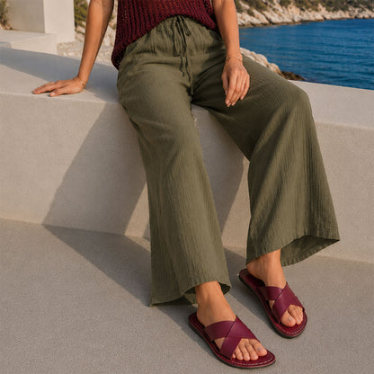 Marsala | Model wearing green pants and red wine-colored leather flat sandals against a coastal beach background
