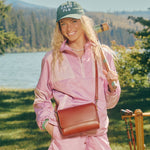 A woman outdoors by a lake wearing a pink tracksuit and green cap with 'CAMP PLG' text, wearing a Madrone Metro Crossbody bag.